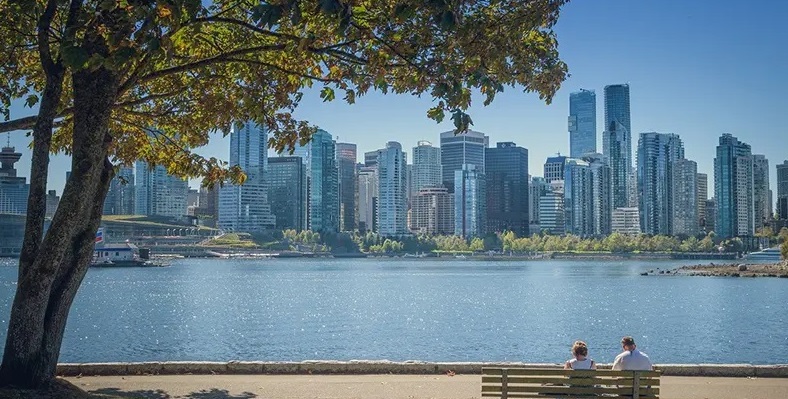 Casal sentado em um banco, com um lago e prédios ao fundo, no Park Stnaley, em Vancouver, no Canadá